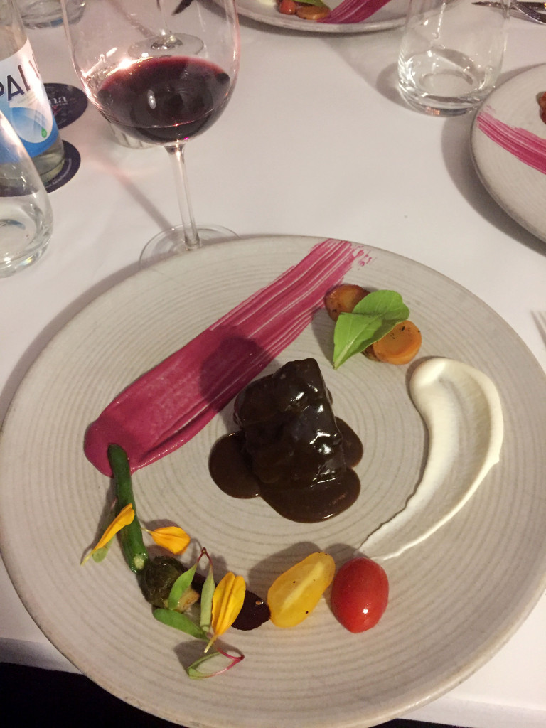 Braised pork with Colombian coffee sugar cane reduction, beet sauce to the left, artichoke heart emulsion to the right, various vegetables and flower petals as garnish.